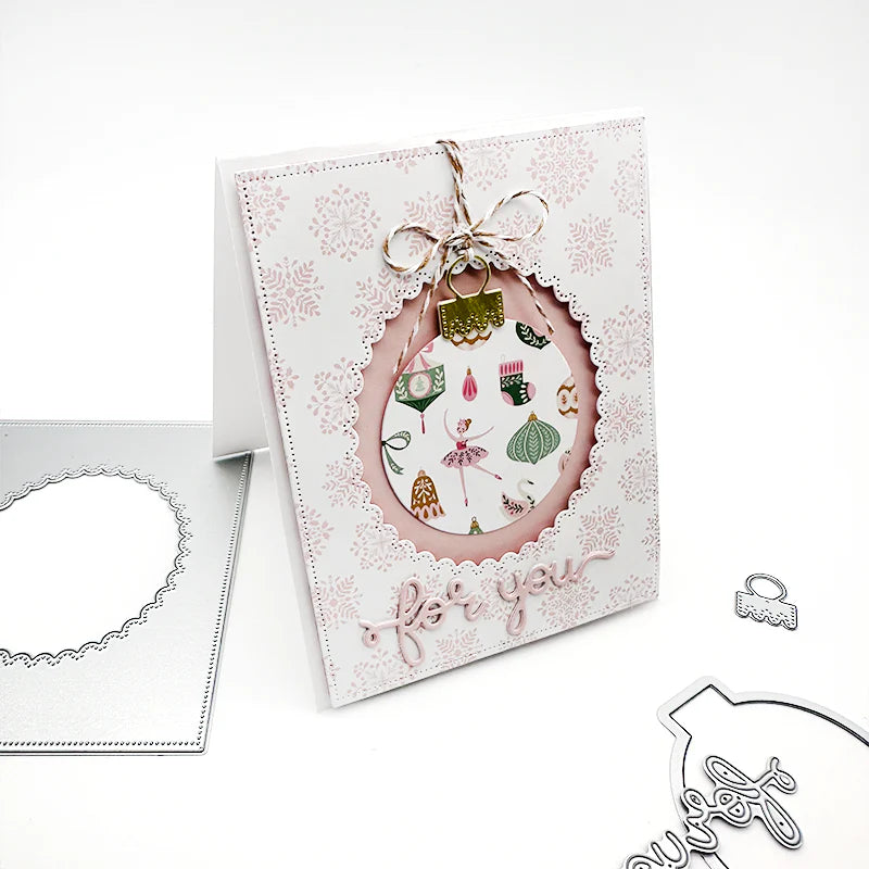 A handmade greeting card with a scalloped white border, a pink inner oval, and a decorative hanging ornament motif, shown standing on a white background with metal die-cutting tools nearby.