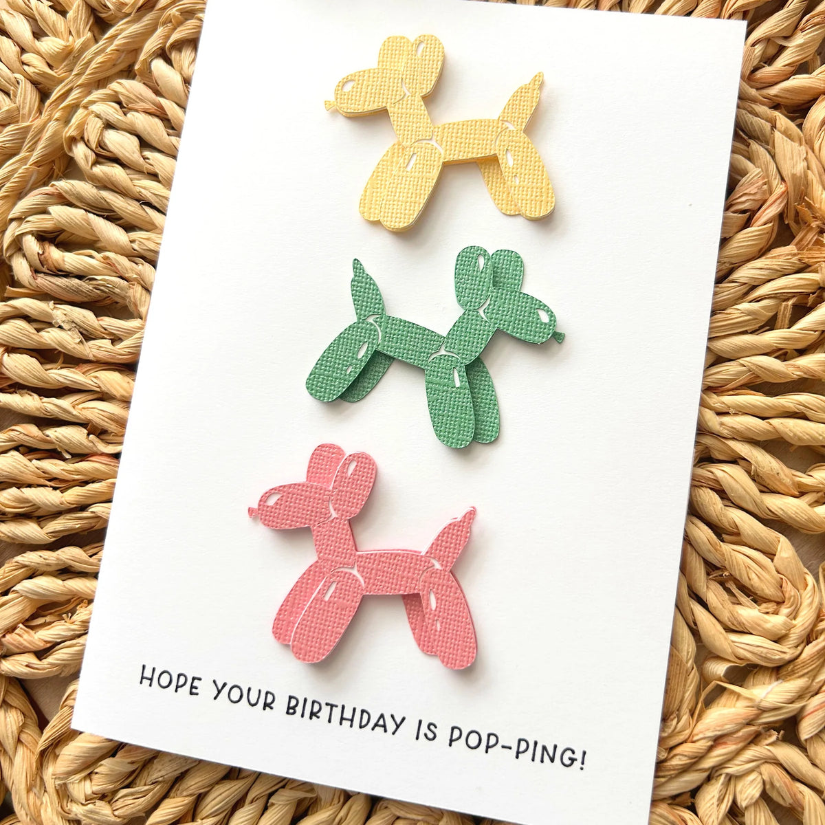 Colorful balloon dog-shaped die cuts on a birthday card with text, placed on a woven surface.