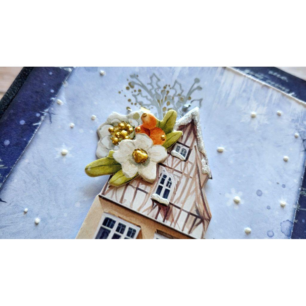 A close-up of a decorative handmade card or art piece featuring a small house with brown timber framing, white flowers with gold centers, green leaves, and a textured light blue background with white dots.
