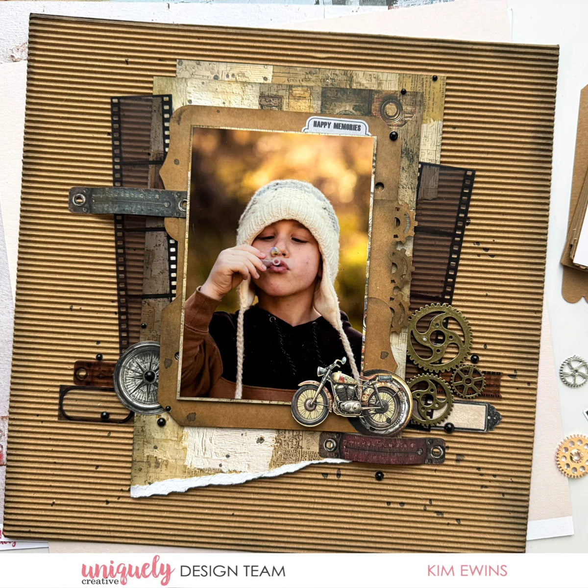 A scrapbook or craft layout featuring a central photo of a child wearing a cream knit hat, surrounded by vintage-style embellishments including gears, a compass, a small motorcycle cutout, torn paper, and metallic accents on a brown corrugated cardstock background. Text at the bottom reads 'uniquely DESIGN TEAM' and 'KIM EWINS'.