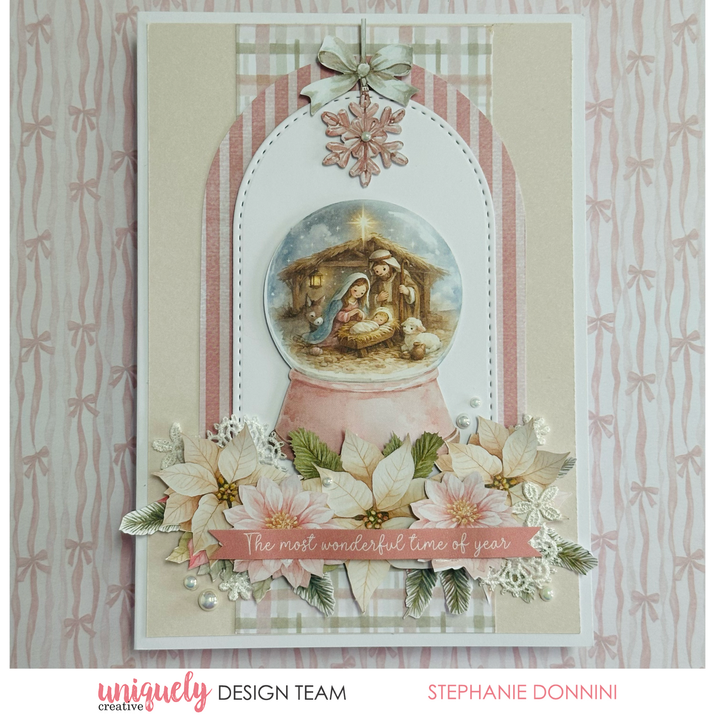 A handmade Christmas greeting card featuring a nativity scene inside a pink snow globe, decorated with poinsettias, white lace, pearls, and a striped arch background. The card is photographed against a patterned pink and white bow backdrop.