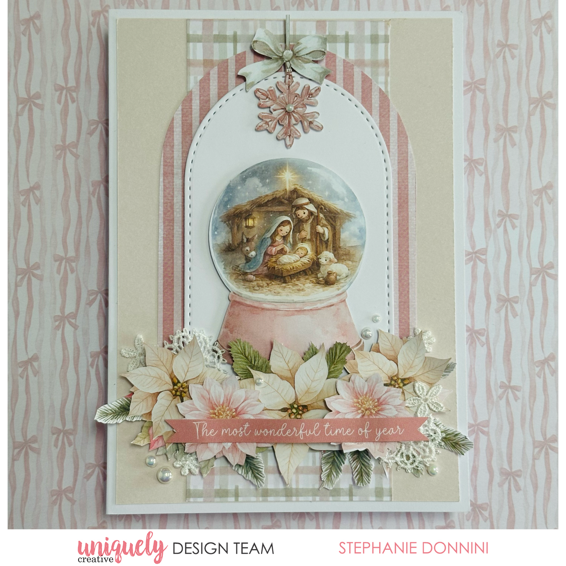 A handmade Christmas greeting card featuring a nativity scene inside a pink snow globe, decorated with poinsettias, white lace, pearls, and a striped arch background. The card is photographed against a patterned pink and white bow backdrop.