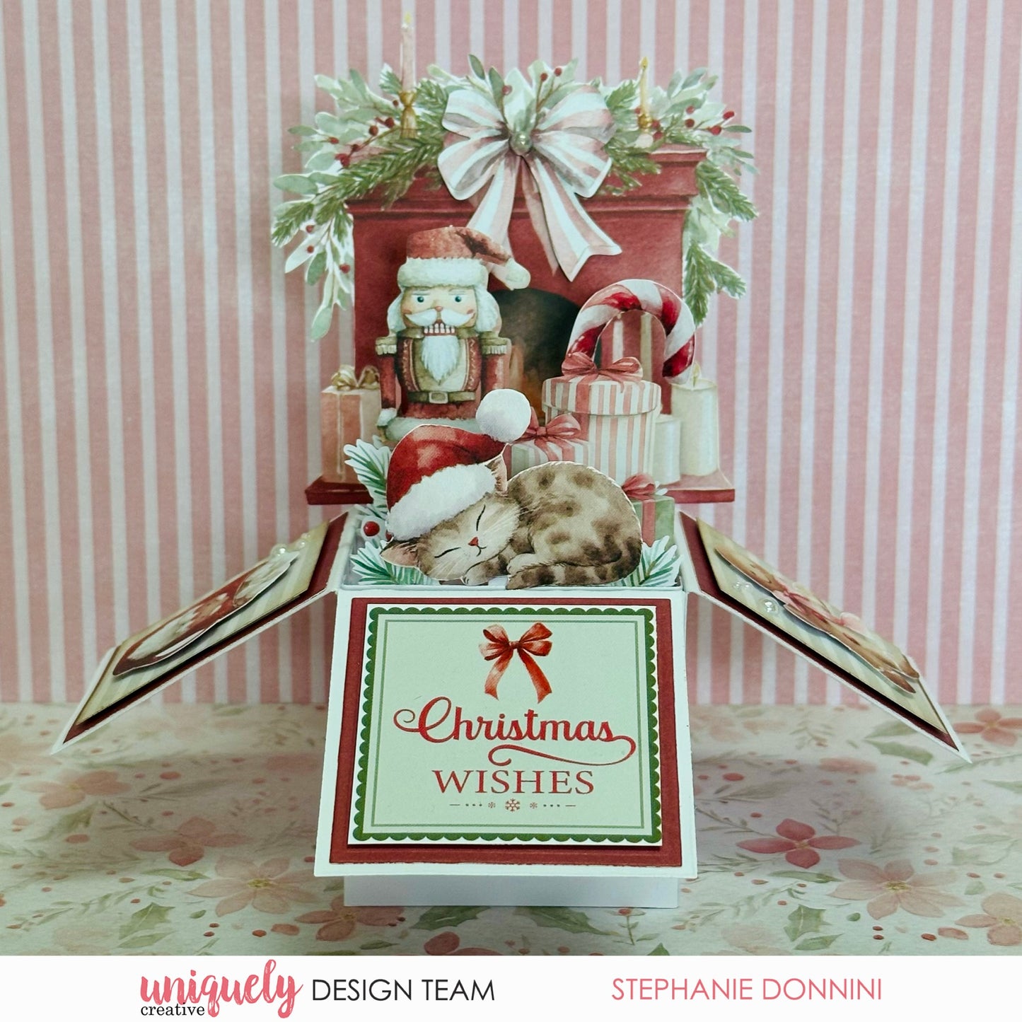 A Christmas-themed 3D pop-up box craft by Stephanie Doninni, featuring a sleeping cat under a nutcracker, candy canes, ornaments, and a large bow, with the front panel reading 'Christmas Wishes'.