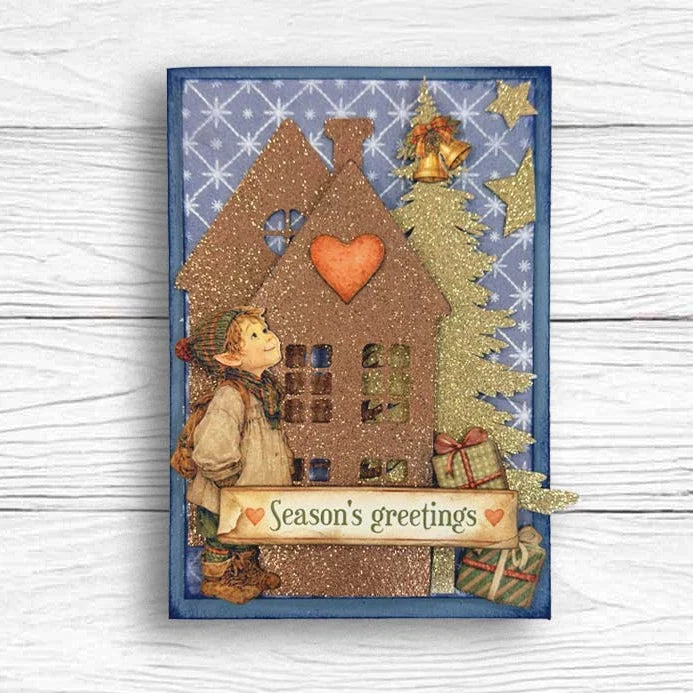 A square holiday greeting card featuring a whimsical house, a child, a gold glitter tree, and a banner reading 'Season's greetings', photographed against a white wood background.