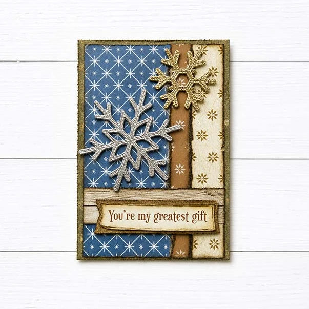 A handmade Christmas greeting card with a winter theme, featuring blue patterned paper, beige patterned paper, brown ribbon accents, glittery snowflakes, and a banner that reads 'You're my greatest gift', displayed on a white wood background.