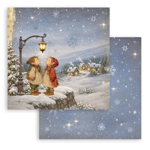 A square winter-themed 12x12 art print featuring two children standing by a snow-covered stone wall under a glowing streetlamp, with snowflakes falling over small houses in the distance. A second matching blue snowflake patterned paper is partially visible behind it, both on a white background.